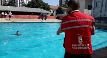 Agente da Defesa Civil realiza treinamento acompanhado por bombeiro. Na imagem, o agente puxa um voluntário que está dentro de uma piscina com uma corda