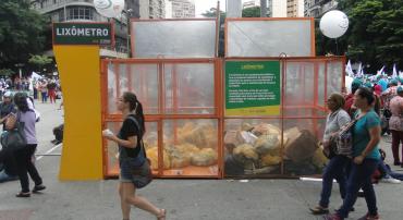 Caixas de material transparente na Praça Sete com lixo ensacado, transeuntes passam ao lado.