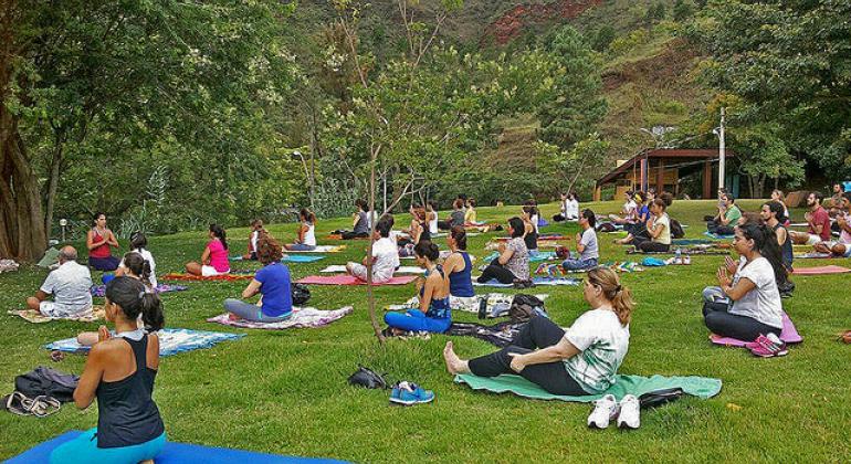 Cerca de 28 pessoas praticam yoga em um parque municipal, ajoelhadas em seus tapetes, durante o dia. 