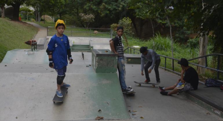 Na foto consta quatro garotos em uma pista de skate, um está sobre o skate de frente para a pessoa que está tirando a foto, outro encostado em uma mureta, um outro no fundo também em cima do skate porém meio agachado e parece que está tentando fazer uma manobra com seu skate, o ultimo garoto está sentado e olhando para o garoto que supostamente está fazendo a manobra. 