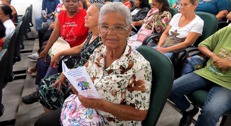 Cerca de 13 idosos sentados em cadeiras, à frente, uma das idosas está de óculos com um panfleto na mão. 