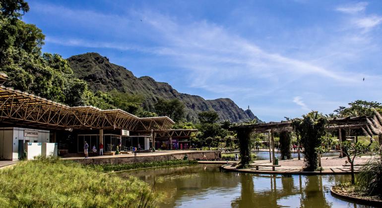 Parque das Mangabeiras com espelho d'água e grama à frente, área administrativa coberta com diversas salas e pessoas e caramanchão à direita. Ao fundo, Serra do Curral sob céu azul de dia. 