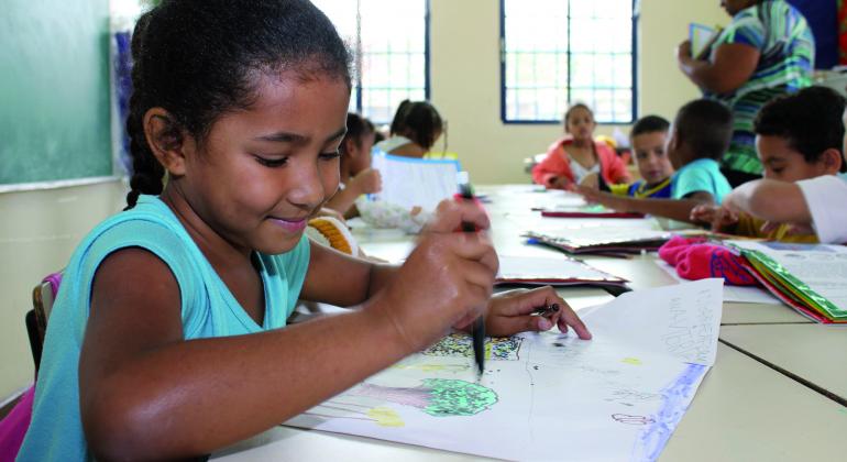 A foto mostra sete crianças em uma sala de aula,com um quadro de escrever verde e duas janelas grandes. À frente v morena, com o cabelo preso em uma trança, vestindo uma camiseta azul clara, segurando um lápis preto, colorindo o desenho de uma árvore. Também vê-se uma professora de costas, segurando alguns papéis.