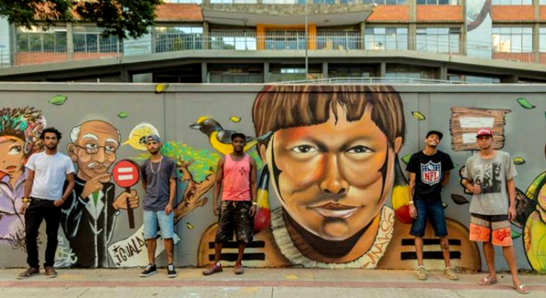 Quatro rapazes de bermuda e boné estão parados à frente de muro grafitado.