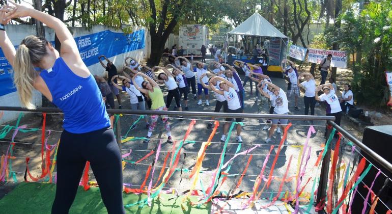 Instrutora de ginástic, em cima de palco, faz movimentos e é acompanhada por mais de vinte pessoas em local com muito verde. 