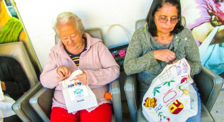 Uma senhora e uma mulher fazem bordados coloridos sentadas em poltronas.