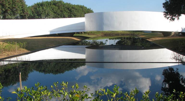 Espelho d'água em frente ao Memorial de Imigração Japonesa. à frente, algumas plantas. Ao fundo, céu azul e árvores.