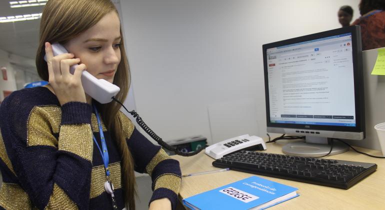 Estagiária sentada à mesa de trabalho atende ao telefone.