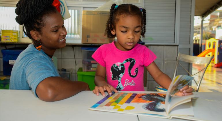 Mulher acompanha criança na leitura de livro infantil.