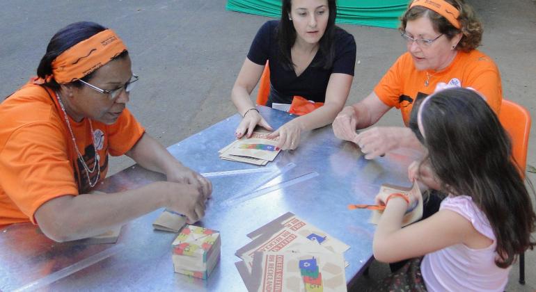 Quatro mulheres de diferentes idades, sentadas em uma mesa, fazendo oficina de reaproveitamento de papel.