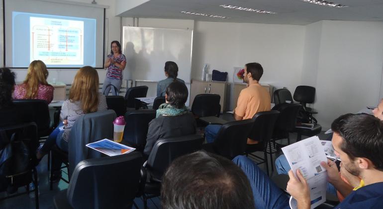 Instrutora da Sudecap em aula expositiva com projeção no quadro branco; nas carteiras, ceca de sete alunos acompanham a exposição.