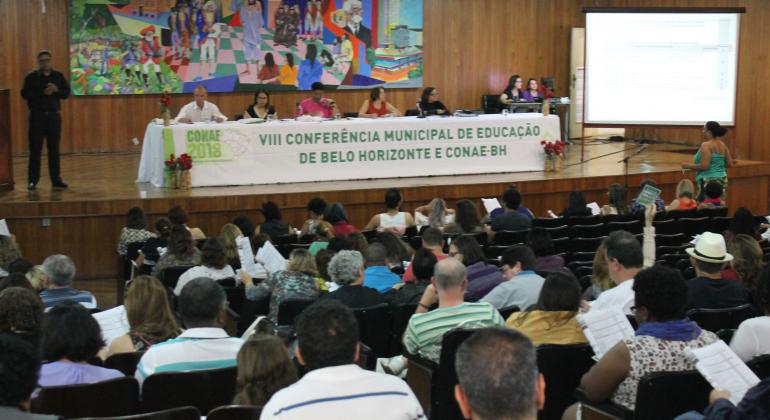 Auditório com cerca de cem pessoas na VIII Conferência Municipal de Educação e Conae- BH. No palco, há uma mesa composta por seis autoridades da educação. Atrás do palco, na parede, vê-se um enorme e colorido painel.