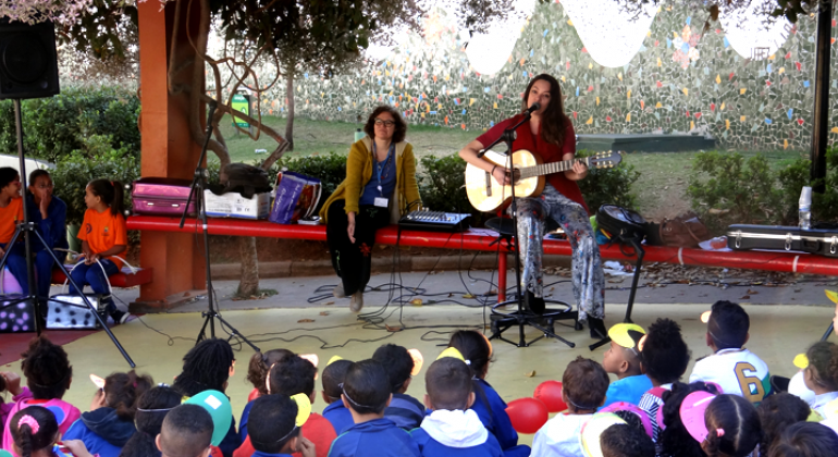 Mulher faz apresentação musical de violão para mais de vinte crianças, ao fundo, outra mulher observa.