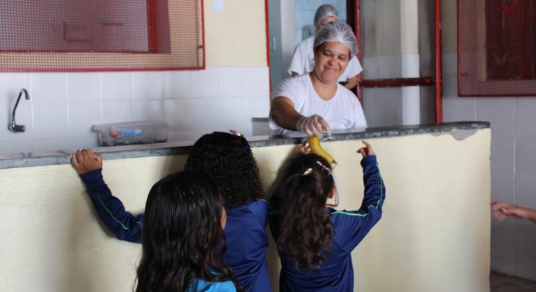 Três meninas na fila para a merenda enquanto uma funcionária entrega uma banana para uma delas