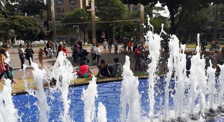 Fonte da savassi, jorra água, ao fundo, cerca de quinze pessoas caminham e aproveitam o dia ensolarado. 