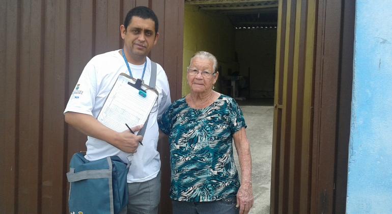 Agente de combate a Endemias em frente a casa de idosa com prancheta na mão e bolsa da Prefeitura.