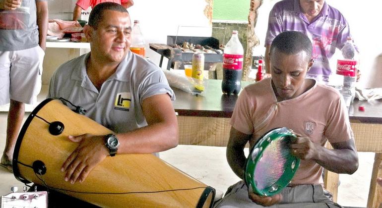 Foto de Gari engenheiro tocando instrumento de percussão ao lado de seu amigo, que toca pandeiro. 