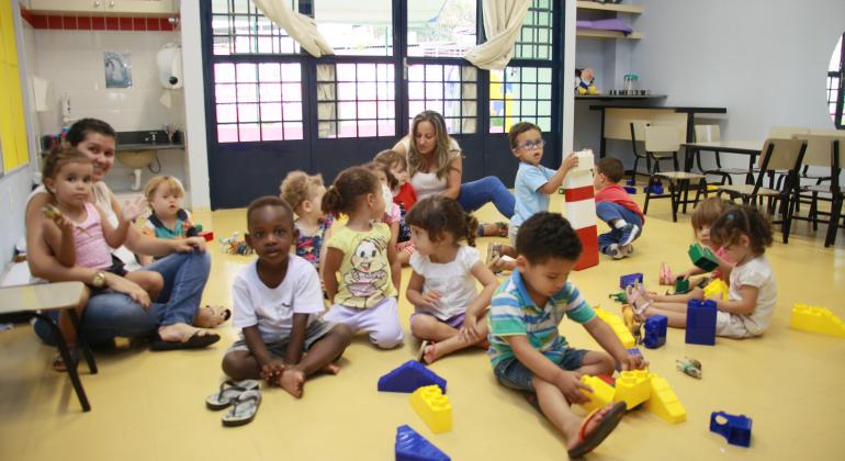 Em sala de aula, cerca de 10 crianças sentadas empilham blocos ao lado de duas professoras 