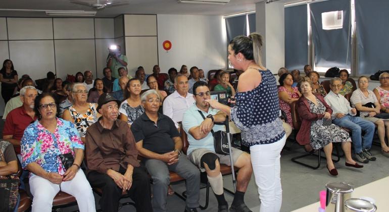 MUlher, de pé, palestra para cerca de trinta pessoas sentadas em sala. 