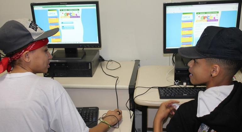 Dois estudantes, cada sentdo um em frente a um computador, se olham. Nas telas, a página do Orçamento Participativo da Criança e do Adolescente (OPCA). 