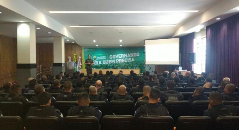 Grupo de cerca de 80 guardas estão sentados em um auditório, de costas para a foto. No palco, de frente, está um membro do exército brasileiro. Um banner escrito "Governando para quem precisa" está ao fundo.