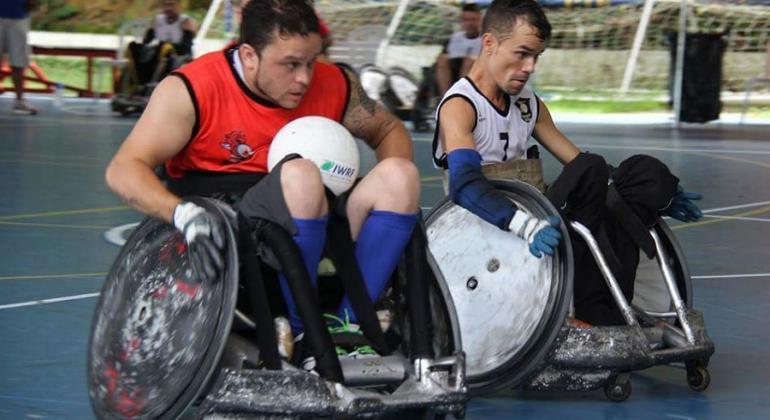 Dois jogadores de rugby em cadeiras de rodas em ação em quadra de esportes. 