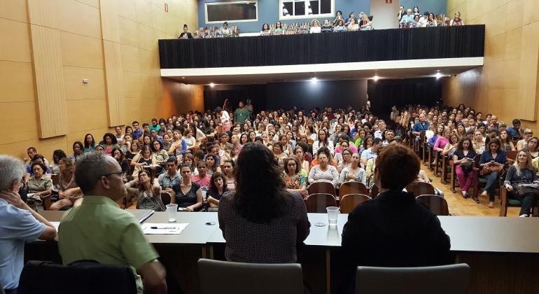 Três pessoas na mesa de costas, em primeiro plano, e atrás o público de mais de cem pessoas lotando o teatro da Faculdade de Medicina da UFMG