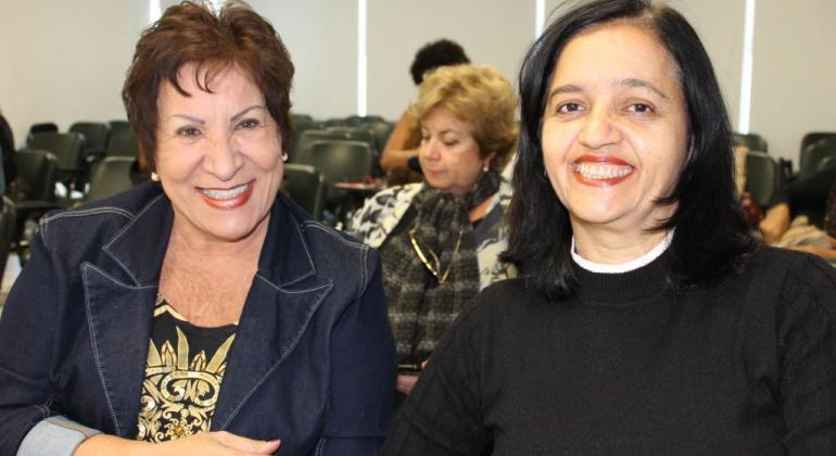 Duas aposentadas da Prefeitura de BH sentadas, sorrido, ao fundo, uma terceira senhora está sentada.