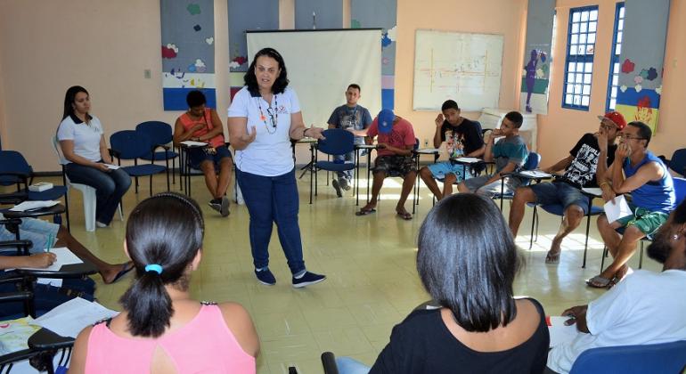 Cerca de treze jovens, sentados e em círculo, assistem a aula de instrutora que está de pé, em uma sala, durante o dia.