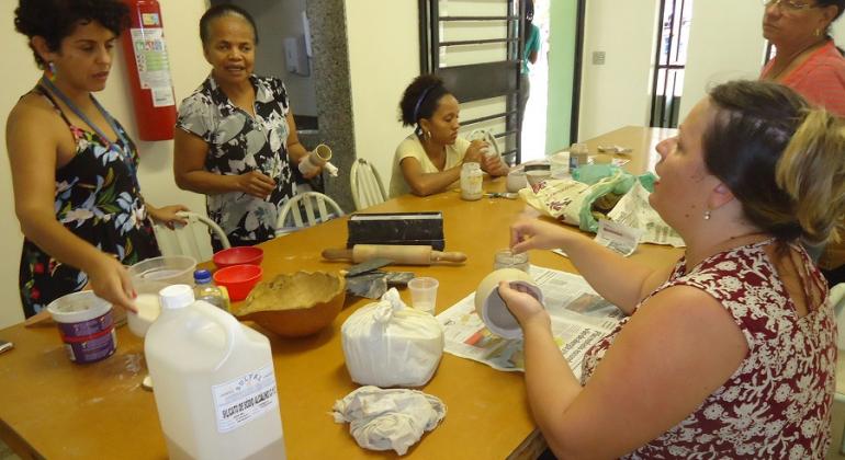 Cinco mulheres pintam cerâmica em torno de uma mesa. 