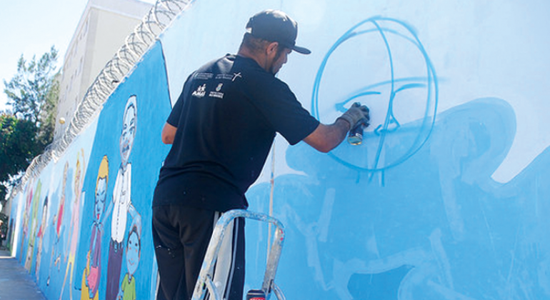 Homem grafita muro em cima da escada, durante o dia.