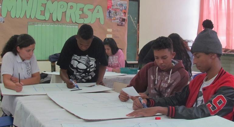 OIto jovens em sala com mesas, sentados, fazendo oficina de artes. 