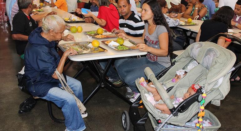 Cidadãos almoçam em restaurante popular, à direita, ao lado de uma mulher, um carrinho dois bebês. 