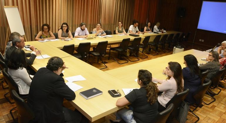 Cerca de treze pessoas, sentadas em uma mesa em forma de "u", conversando. Ao fundo, um telão. 