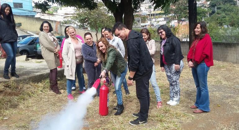 Cerca de nove pessoas participam de treinamento preventivo para para utilização de extintores em local aberto; mulher aciona extintor acompanhada de instrutor.