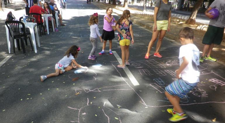 Cinco crianças desenham na rua com giz e brincam, no fundo, dois aultos sentados acompanham. 