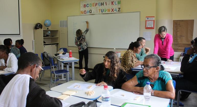 Seis alunos adultos em sala de aula, duas professoras orientadoras acompanham, uma no quadro branco, outra acompanhando o aluno