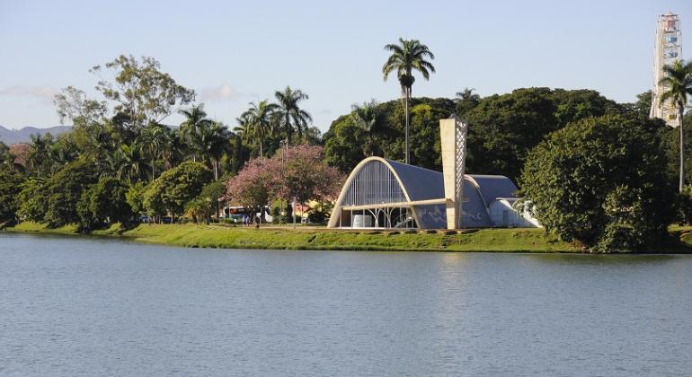 Igreginha da Lagoa da Pampulha atrás da Lagoa. Ao fundo, muitas árvores e um céu azul