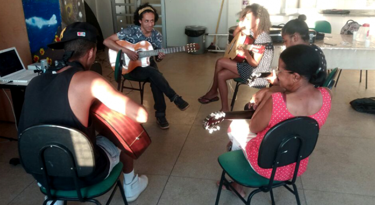 Seis pessoas, sentadas em uma sala, fazem oficina de violão. 