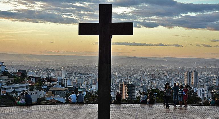 Cruz da Praça do Papa à frente; ao fundo, cidadãos vêem a cidade de BH com muitos prédios e o horizonte. 