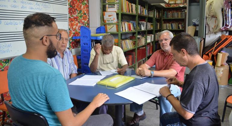 Cinco pessoas sentadas em uma mesa com cadernos; ao fundo, vários livros em uma estante.