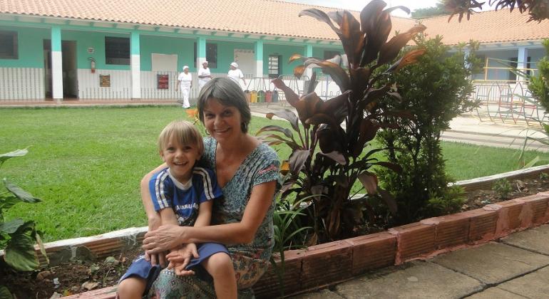 Mãe abraça filho sentada em canteiro com plantas, ao fundo, gramado e uma casa. 