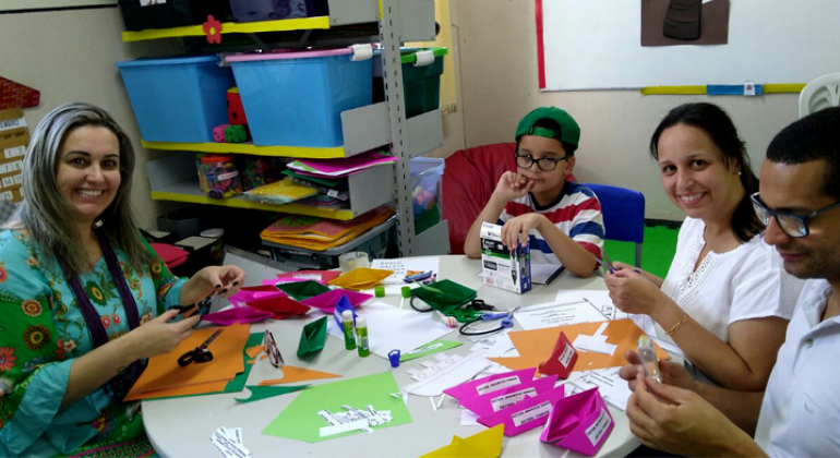 Pais participam com o aluno de atividades propostas pela professora na sala de Atendimento Educacional Especializado; sentados à mesa, manipulam papéis coloridos e cola em bastão.