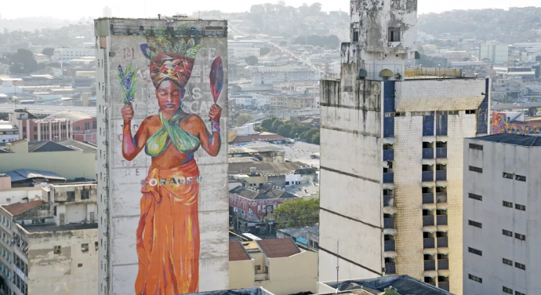 Mulher afro com turbante na cabeça grafitada em lateral de edifício. Em sua cintura, os dizer "coragem". Ao fundo, mais prédios e cenário urbano.