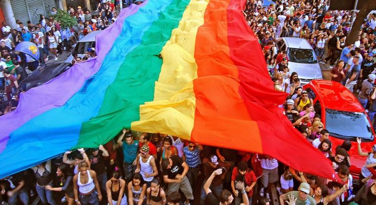 Mais de cem pessoas na rua segurando uma enorme bandeira nas cores do arco-íris, símbolo da luta LGBT. 