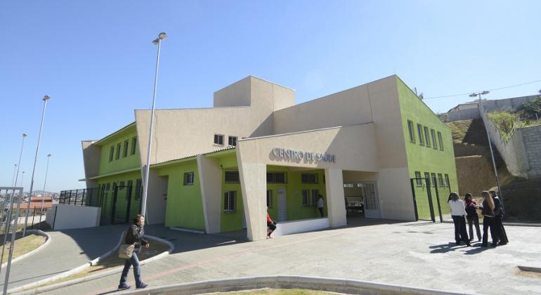 Fachada de centro de saúde, com quatro cidadãos transitando na calçada, durante o dia.