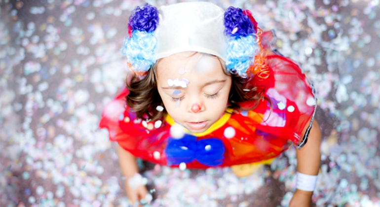 Criança fantasiada de palhaço fecha os olhos; ela está em meio a confetes de papel. 
