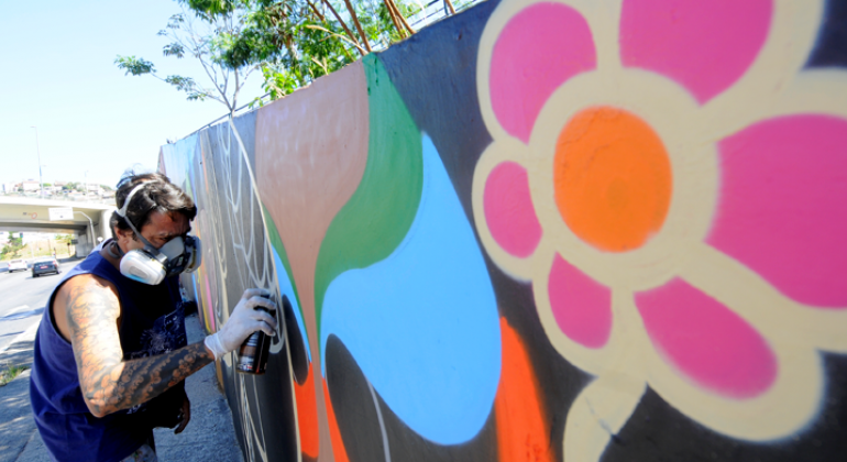 Homem com máscara de segurança e luva grafita muro com flores e cores vibrantes, durante o dia.