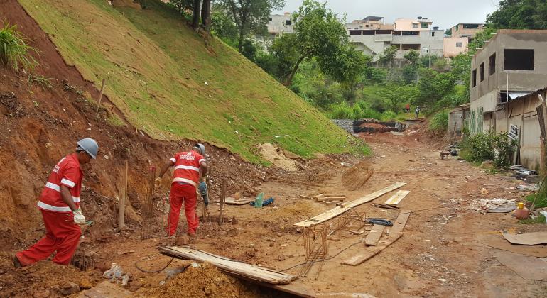 Dois trabalhadores com uniformes da Prefeitura executam obra em encosta no Barreiro. Ao lado, há construções inacabadas e a rua está sem capeamento.