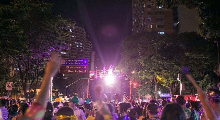 Bloco Chama o Síndico com caminhão elétrico e prédio ao fundo, muitas pessoas à frente e placas indicativas de trânsito à direita. 